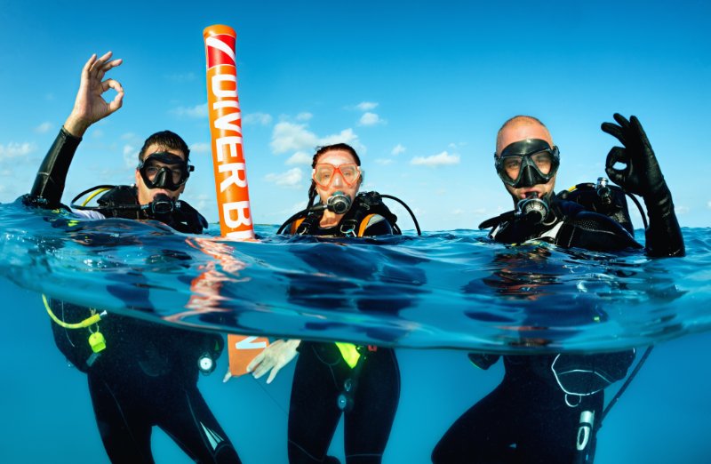 Three scuba divers floating above the water showing the OK hand symbol with their thumb and index fingers