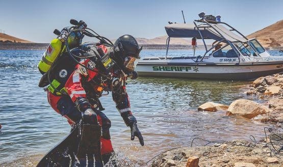 A rescue diver in full gear on the shore with a boat in the background