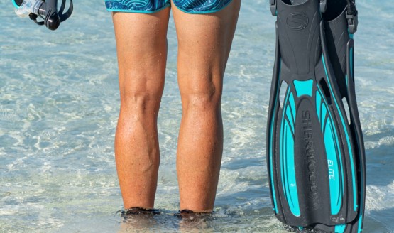 A man on the beach looking out over the water with a pair of snorkeling fins in one hand and a mask in the other.