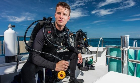 A male scuba diver on a boat, he's wearing his full scuba gear, but his face is visible.