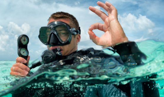 A diver above water holding an analog pressure gauge. He's making the OK symbol with his thumb and index finger