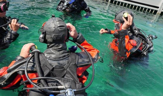 Four public safety divers in drysuits. They are standing in water and adjusting their gear.