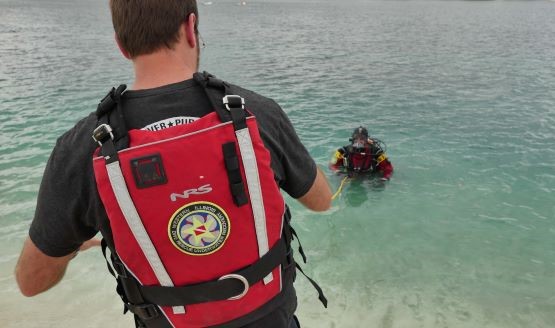 A dive instructor is standing onshore and watching a public safety diver in open water.