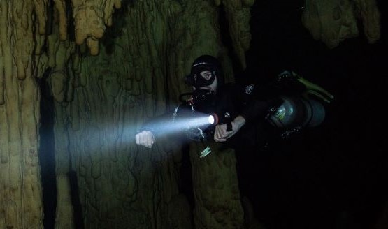 A diver shining his dive light in front of him. There's a cavern wall behind him. width=