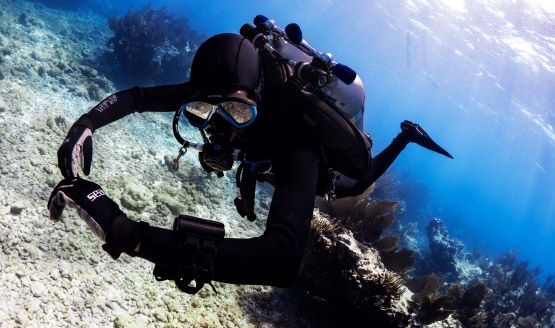 A technical diver clad in a black wetsuit and hood, underwater, and looking at his wrist-mounted dive computer