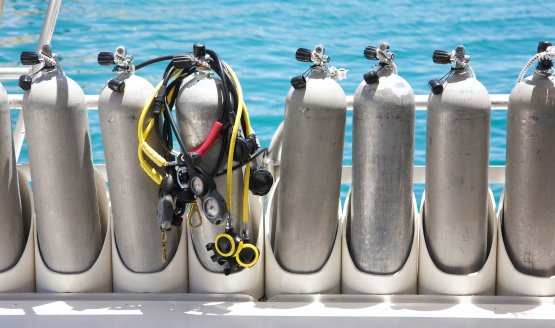 Eight scuba tanks lined up in a row on a dock with water behind them.