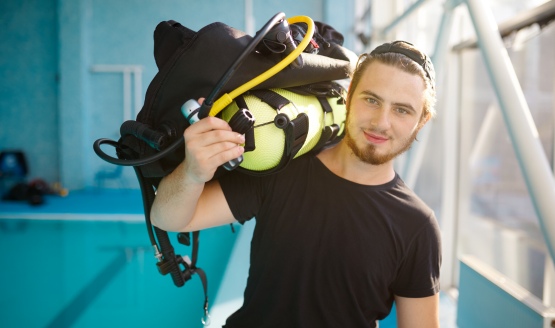 A diver standing near an indoor pool with scuba gear resting on his shoulder.